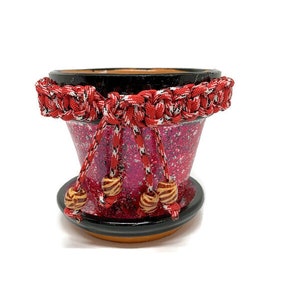 May include: A red and black terracotta planter with a red braided rope trim and wooden beads. The planter has a speckled pink and white glaze.