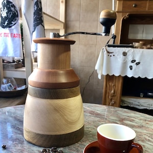May include: A wooden coffee grinder with a hand crank and a brown ceramic coffee cup on a white saucer. Coffee beans are scattered on the table.