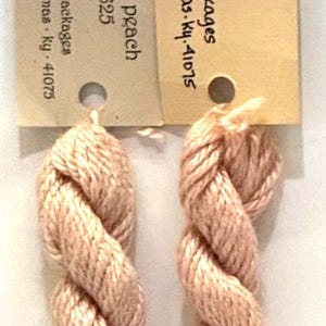 May include: Two skeins of peach-colored yarn, tightly twisted and displayed vertically. The yarn is a soft, light shade of peach. Each skein is attached to a small tag with text. The yarn is likely for crafts or knitting.