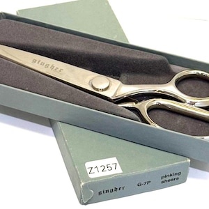 May include: Silver-toned Gingher pinking shears in a gray box with a dark gray interior. The shears are in a partially open box, with the brand name "gingher" engraved on the blade. The box is labeled "Z1257" and "G-7P pinking shears."