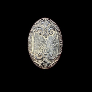 May include: An oval, antique-style metal decorative element with ornate floral and scrollwork designs. The central area has a textured surface. The piece is in shades of silver and bronze against a black background.