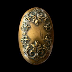 May include: Oval-shaped antique brass door knob with ornate floral and leaf design. The knob has a slightly tarnished finish, with detailed relief carvings. The background is solid black.