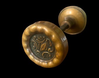May include: Antique brass door knob with an oval face featuring a floral design. The knob has a decorative, scalloped edge and a round handle. The metal has a warm, aged patina, suggesting vintage craftsmanship. The background is black.