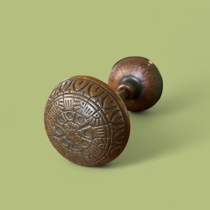 May include: Two antique bronze door knobs. The front knob has a detailed, circular pattern. The second knob is in the background. The knobs are on a light green background. The knobs are likely for a door or cabinet.