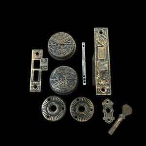 May include: An assortment of antique brass door hardware components, including two round doorknobs with intricate patterns, a rectangular faceplate, and various other pieces. The hardware has a vintage aesthetic with a dark, aged finish.