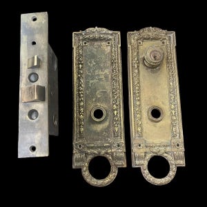 May include: Antique brass door hardware set with ornate details. The set includes a door knob, a door plate, and a lock. The door knob has a keyhole and a round handle. The door plate has a decorative border and a keyhole. The lock has a lever handle and a keyhole.