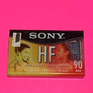 May include: A vintage Sony HF 90 min audio cassette tape. The cassette features the Sony logo and the text "HF HIGH FIDELITY". The packaging shows images of people listening to music. The cassette is on a pink background.
