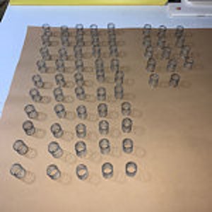 May include: A collection of small, clear, cylindrical objects arranged on a light brown surface. The objects cast shadows, suggesting a light source. The arrangement appears organized, with the objects standing upright.