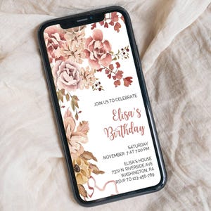 Könnte beinhalten: Ein Smartphone zeigt eine florale Geburtstagseinladung. Die Einladung zeigt ein Aquarelldesign mit rosa, pfirsichfarbenen und braunen Blumen. Der Text lautet "Elisa's Birthday" mit Veranstaltungsdetails. Das Telefon liegt auf einem beigen Stoff.