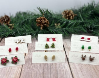May include: A collection of Christmas-themed stud earrings, including gingerbread men, Christmas trees, Santa hats, and reindeer. The earrings are displayed on small cards against a backdrop of pine branches and pinecones.