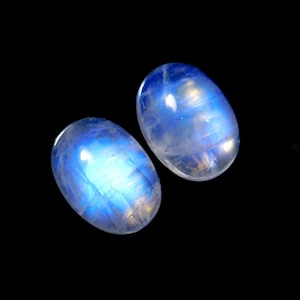May include: Two oval-shaped moonstone cabochons featuring a translucent appearance with a distinct blue adularescence or glow. The gemstones have a smooth, polished surface and exhibit internal inclusions and light reflections against a black background.