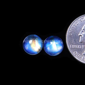 May include: Two round, blue gemstones with a quarter coin for size comparison. The gemstones have a luminous, iridescent quality with hints of yellow and orange. The background is solid black, highlighting the gems' color and shape.