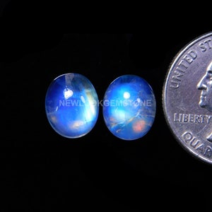 May include: Two oval-shaped moonstone cabochons, approximately 10mm in size, with a blue and iridescent sheen. The stones are displayed next to a US quarter for size comparison.
