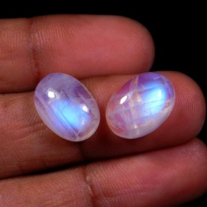 May include: Two oval-shaped moonstone cabochons with a iridescent blue sheen. The stones are a pale white color with a subtle pink hue.