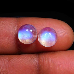 May include: Two round, cabochon-cut moonstones featuring a translucent appearance with a distinct blue adularescence or glow. The stones are smooth and polished, displaying a soft iridescence across their surfaces. The text NEWLOOKGEMSTONE is visible.