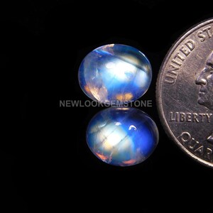 May include: A pair of oval-shaped moonstone cabochons, one of which is being compared to a US quarter for size. The moonstones are iridescent with a blue and white sheen.