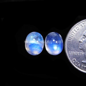 May include: Two oval-shaped moonstone cabochons, approximately 1/2 inch in size, with a blue iridescent sheen. The stones are displayed next to a US quarter for size comparison.