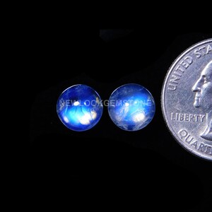 May include: Two round, blue gemstones with a quarter coin for size comparison. The gemstones have a luminous quality, with white and blue internal patterns. The background is black, highlighting the gems' color and shape.