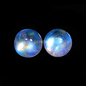 May include: Two round, cabochon-cut moonstones featuring a translucent, milky white base with a vibrant blue adularescence and internal inclusions. The text NEWLOOKGEMSTONE is overlaid across the center of both stones against a black background.