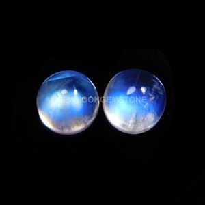 May include: Two round, cabochon-cut moonstones featuring a translucent appearance with a distinct blue adularescence glow. The stones display internal inclusions and the text NEWLOOKGEMSTONE is overlaid across the center of both items.