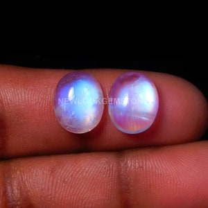 May include: Two oval-shaped moonstone cabochons with iridescent blue and purple colors. The stones are smooth and polished.
