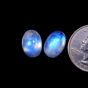 May include: Two oval-shaped, polished moonstone gemstones with a blue and white iridescent glow. The gemstones are displayed on a black background next to a quarter for size comparison. The stones are likely for jewelry making.