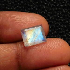 May include: A square-shaped, iridescent moonstone gemstone with a light blue and yellow sheen.