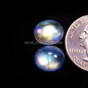 May include: Two oval-shaped iridescent moonstone cabochons, one with a blue and yellow sheen and the other with a blue and pink sheen, next to a US quarter for size comparison.