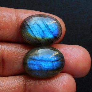 Puede incluir: Dos piedras preciosas de labradorita de forma ovalada. Las piedras muestran un vibrante juego de colores, con azules y verdes iridiscentes arremolinándose en sus superficies. Las piedras pulidas se colocan sobre un fondo oscuro, resaltando su belleza natural.