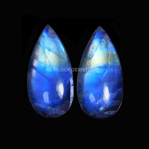 May include: Two pear-shaped moonstones featuring a translucent appearance with a vibrant blue adularescence and internal inclusions. The text NEWLOOKGEMSTONE is centered between the two gemstones against a solid black background.