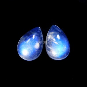 May include: Two pear-shaped moonstone cabochons featuring a translucent body with a vibrant blue adularescence glow. The stones display internal inclusions and a smooth, polished surface. The text NEWLOOKGEMSTONE is visible across the center.
