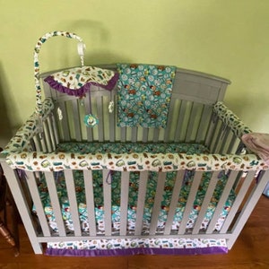 May include: A gray crib with a white and green patterned bedding set. The bedding includes a fitted sheet, a bumper, and a crib skirt. The crib also has a mobile with a purple and white fabric canopy.