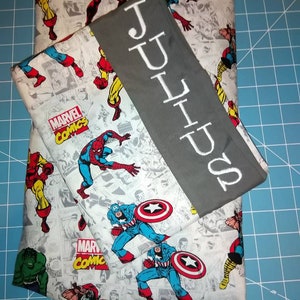 May include: A gray and white fabric with a Marvel Comics theme featuring characters like Spider-Man, Captain America, and Iron Man. The fabric is folded in half and has a gray fabric strip with the name "JULIUS" embroidered in white.