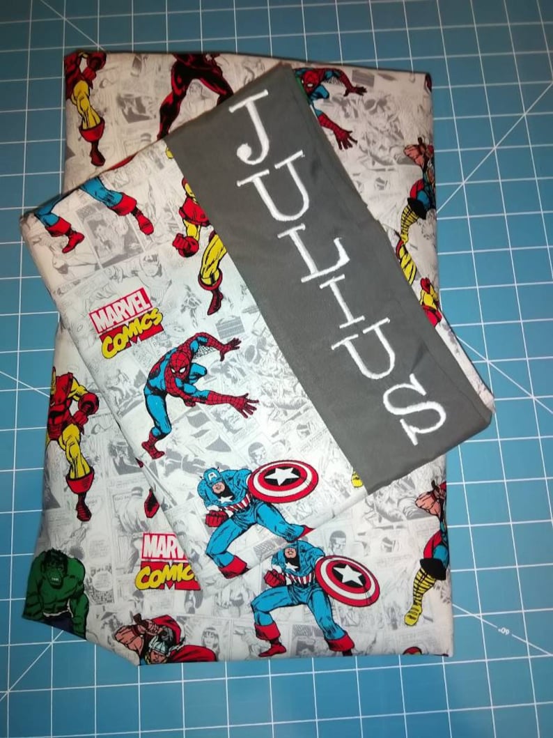 May include: A gray and white fabric with a Marvel Comics theme featuring characters like Spider-Man, Captain America, and Iron Man. The fabric is folded in half and has a gray fabric strip with the name "JULIUS" embroidered in white.