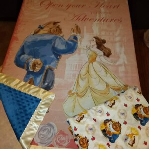 May include: A pink and white blanket with a printed image of Belle and the Beast from Disney's Beauty and the Beast. The text "Open your heart to the adventures" is printed on the blanket. The blanket is made of two layers, a top layer with the printed image and a bottom layer with a blue and white polka dot pattern. The blanket is trimmed with a gold satin ribbon.