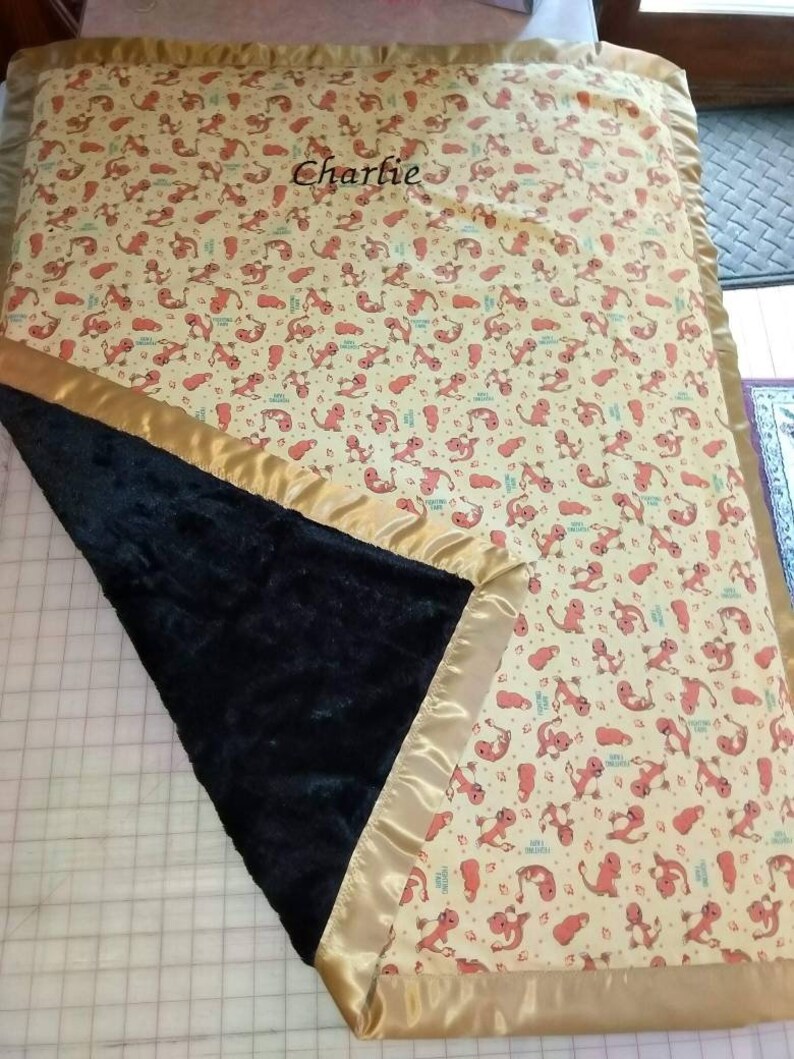 May include: A yellow and orange baby blanket with a black minky backing and gold satin trim. The blanket has a repeating pattern of cartoon lizards and the name "Charlie" embroidered in the center.
