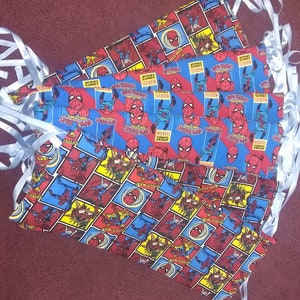 Op de afbeelding: Een stapel blauwe en rode stoffen mondmaskers met een herhalend patroon van Spider-Man-comicboekomslagen. De maskers hebben witte bandjes.