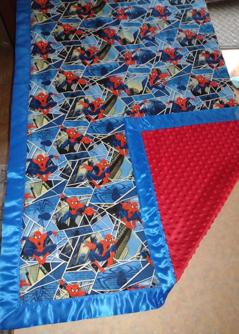 May include: A blue and red baby blanket with a Spiderman pattern on one side and a red minky fabric on the other side. The blanket has a blue satin trim.