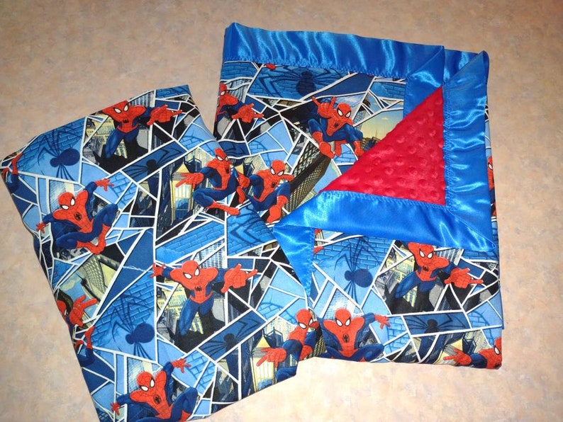 May include: A blue and red baby blanket with a Spiderman pattern. The blanket has a satin border and a soft, textured lining.