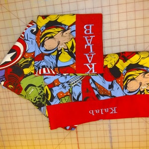 May include: Two red fabric panels with white embroidered names "Kailab" and "Batman". The panels are attached to a multi-colored fabric with a comic book pattern featuring superheroes like Wolverine, Hulk, and Captain America.