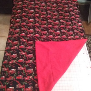 May include: A red and black fleece blanket with a Cars print featuring Lightning McQueen. The blanket has a red border and the name "Logan" is embroidered in the top corner.