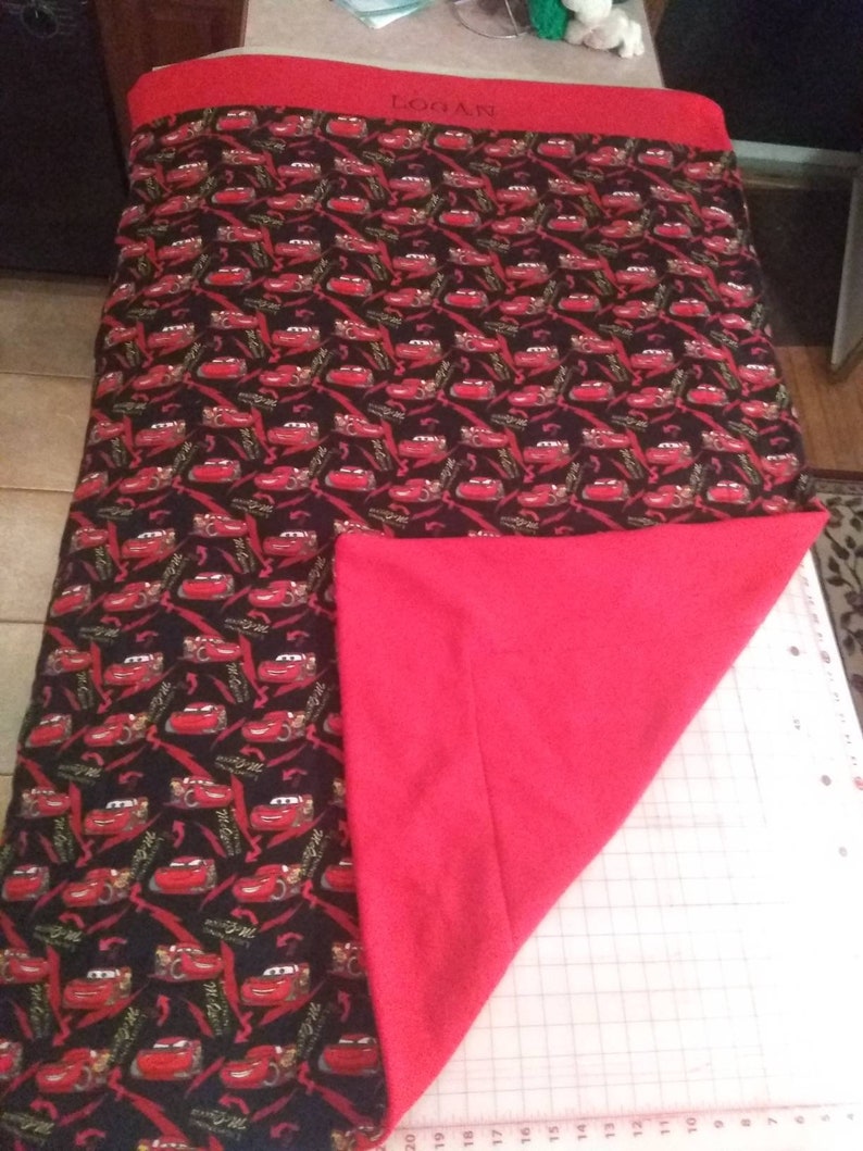 May include: A red and black fleece blanket with a Cars print featuring Lightning McQueen. The blanket has a red border and the name "Logan" is embroidered in the top corner.