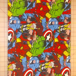 Op de afbeelding: Een kleurrijke stof met een herhalend patroon van verschillende superhelden, waaronder Captain America, Thor, Hulk en Iron Man. De stof is rood, blauw, groen, geel en zwart.