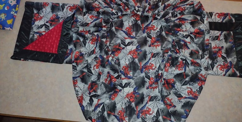 May include: A black and red baby blanket with a Spiderman pattern. The blanket has a soft, plush texture and is perfect for cuddling.