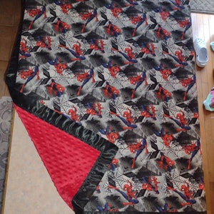 May include: A red and black baby blanket with a Spiderman pattern. The blanket has a soft, plush texture and a satin trim.