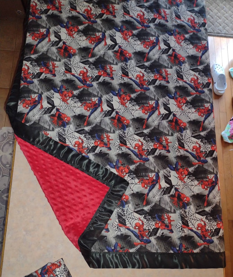 May include: A red and black baby blanket with a Spiderman pattern. The blanket has a soft, plush texture and a satin trim.