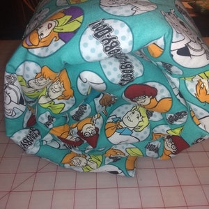 May include: A teal blue fabric with a repeating pattern of cartoon characters from Scooby-Doo. The fabric features the text "Scooby-Dooby-Doo" in a white circle.