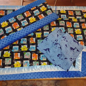 May include: A baby blanket set featuring a black fabric with colorful cartoon characters. The set includes a blanket, a bumper, and two smaller blankets. The set has blue accents and trim, with a soft, textured fabric.