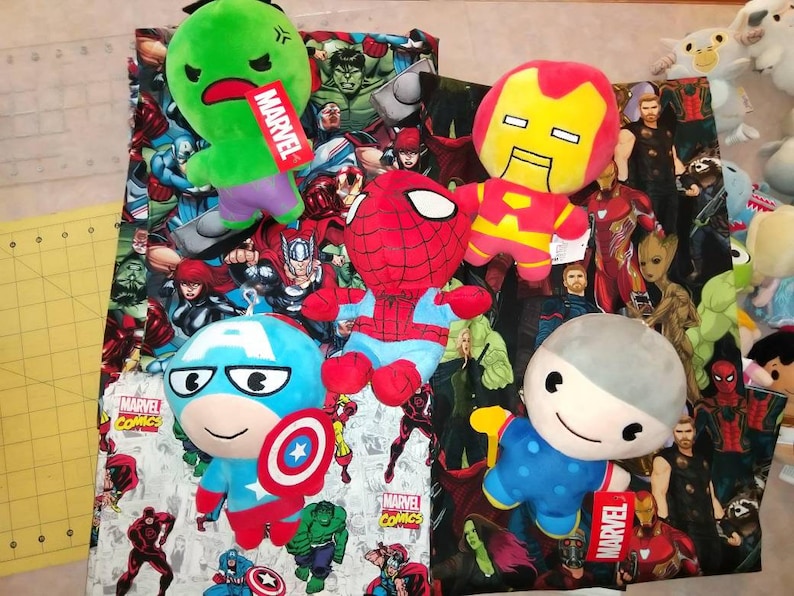 May include: A collection of plush toys featuring Marvel characters. The toys are arranged on a fabric background with a Marvel Comics print. The toys include Iron Man, Spider-Man, Captain America, Hulk, and a grey character with a blue shirt. The fabric background features a variety of Marvel characters, including Iron Man, Spider-Man, Captain America, Hulk, Thor, and Groot.