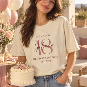 May include: A cream-colored short-sleeve t-shirt featuring the text "CHAPTER 18", the name "Juliana" in script, and the phrases "THE STORY CONTINUES" and "EST. 2008" printed in dark brown and muted pink on the front chest area.